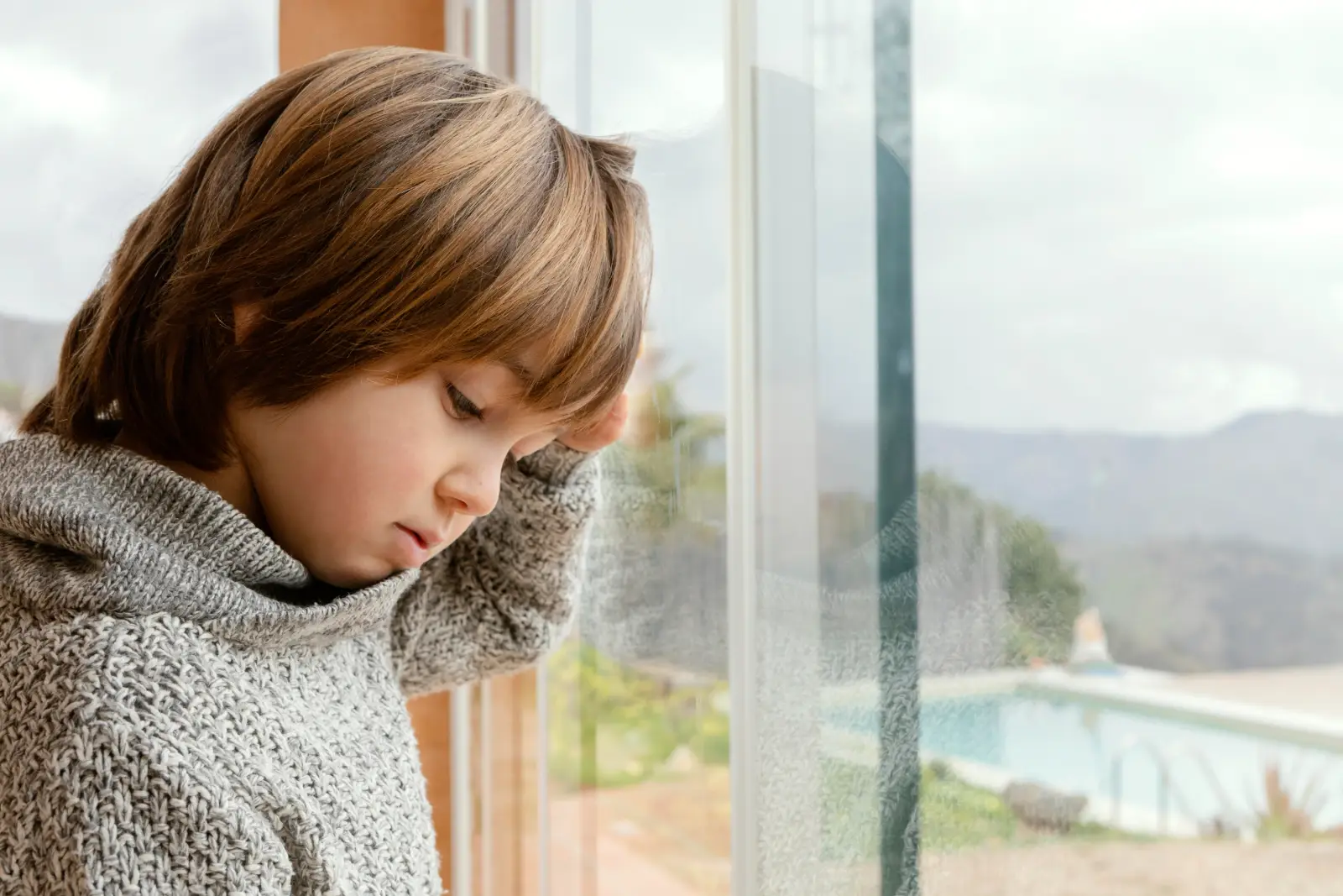 Bored boy standing by the window waiting for the rain to stop